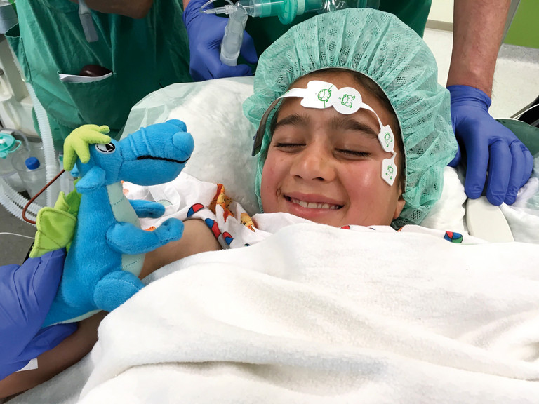 Eine junge Patientin mit einer grünen OP-Haube liegt beim Projekt "Herzbrücke" von Albertinen-Stiftung und Albertinen Herz- und Gefäßzentrzum auf einem Operationstisch und blickt auf einen blauen Stoff-Dinosaurier Eine junge Patientin mit einer grünen OP-Haube liegt beim Projekt "Herzbrücke" von Albertinen-Stiftung und Albertinen Herz- und Gefäßzentrzum auf einem Operationstisch und blickt auf einen blauen Stoff-Dinosaurier