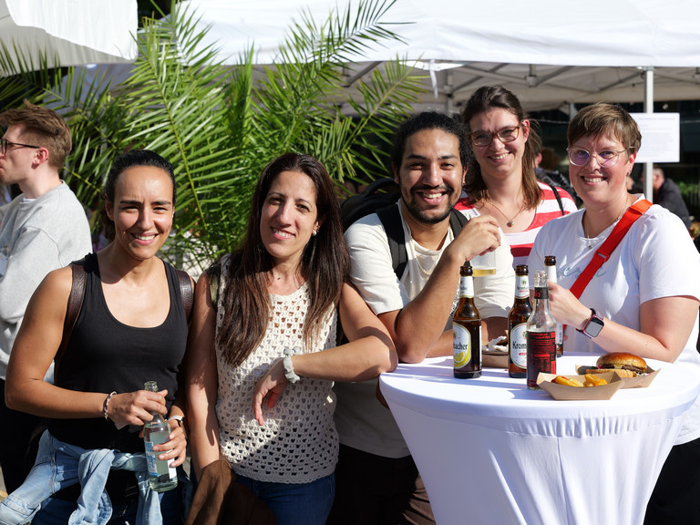 Mitarbeitende an einem weiß gedeckten Stehtisch mit Getränken und Burgern blicken lachend in die Kamera beim Albertinen-Sommerfest für die Mitarbeitenden aus den unterschiedlichen Einrichtungen der Immanuel Albertinen Diakonie am Standort Schnelsen auf dem Platz vor der Kirche am Albertinen Krankenhaus. Mitarbeitende an einem weiß gedeckten Stehtisch mit Getränken und Burgern blicken lachend in die Kamera beim Albertinen-Sommerfest für die Mitarbeitenden aus den unterschiedlichen Einrichtungen der Immanuel Albertinen Diakonie am Standort Schnelsen auf dem Platz vor der Kirche am Albertinen Krankenhaus.
